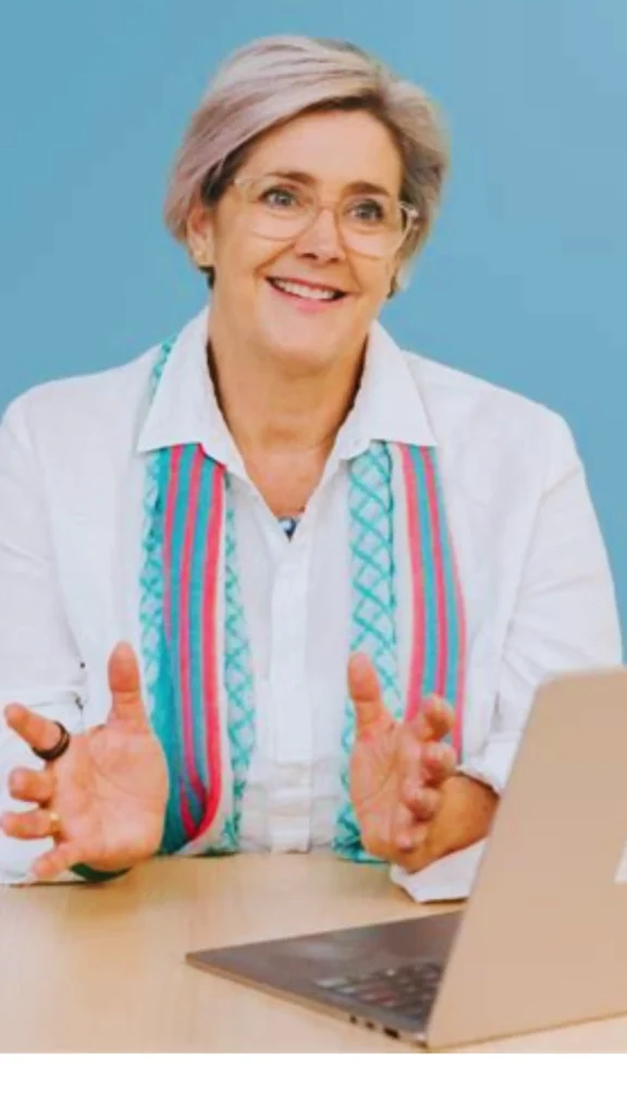 Pinterest pin titled "_Audit What You Already Own.webp". The image features a smiling woman wearing a classic white shirt and a colorful scarf, sitting at a desk with a laptop. This pin serves as an informative guide on the first essential step of building a Capsule Wardrobe for women's fashion: auditing your current closet. It encourages an intentional review of what you already own to simplify your collection and streamline your style effectively.