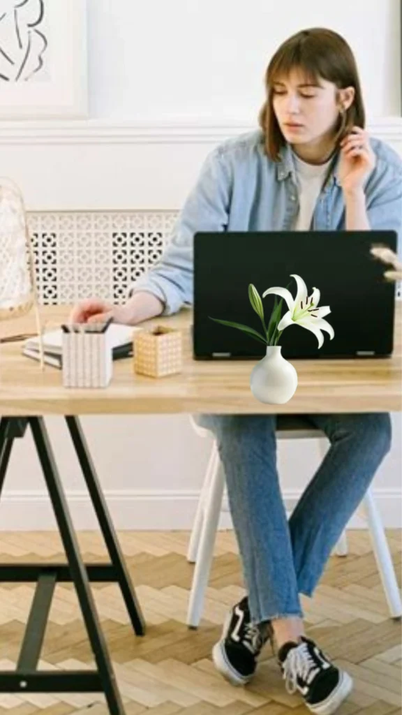 Pinterest pin titled "What If Minimalism Feels Hard.jpg". The image features a woman working at a wooden desk with a laptop and a small vase of lilies. She is dressed in a casual, minimalist outfit of a light denim shirt, jeans, and sneakers. This pin explores the practical realities of adopting a simplified lifestyle, offering support and encouragement for women who may find the transition to a Capsule Wardrobe and minimalist fashion challenging at first.