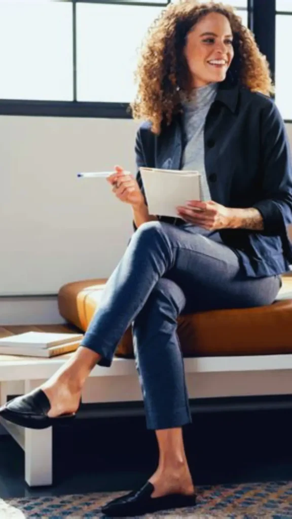 Women's fashion Pinterest pin featuring a smiling woman with curly hair, sitting and taking notes in a bright office space. She is dressed in a professional, minimalist outfit consisting of a grey turtleneck, a dark navy jacket, tailored trousers, and black loafers. This stylish and comfortable look serves as perfect inspiration for a professional Capsule Wardrobe, demonstrating how to combine timeless, versatile pieces for a polished everyday work ensemble.