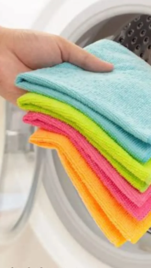 A close-up shot showing a hand holding a stack of colorful, folded microfiber cloths—in shades of light blue, lime green, pink, and orange—in front of an open washing machine. This Pinterest pin highlights the practical and organized side of maintaining a clean and functional home, which is a key aspect of managing your Capsule Wardrobe. By keeping your essentials well-cared for, you ensure your clothing stays in top condition for longer.
