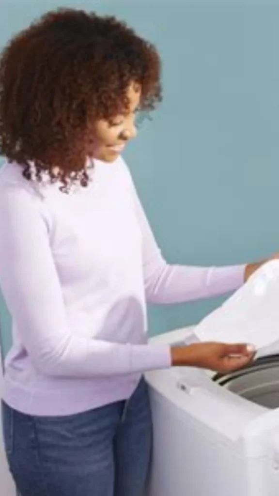 Women's fashion Pinterest pin titled "Your Laundry Schedule Will Completely Change.webp". The image shows a woman with curly hair wearing a light-colored long-sleeve top and jeans, happily loading clothes into a washing machine. This pin highlights how adopting a Capsule Wardrobe can simplify your daily routine, including reducing the frequency of laundry by curating a collection of versatile, high-quality, and easy-to-care-for clothing essentials.