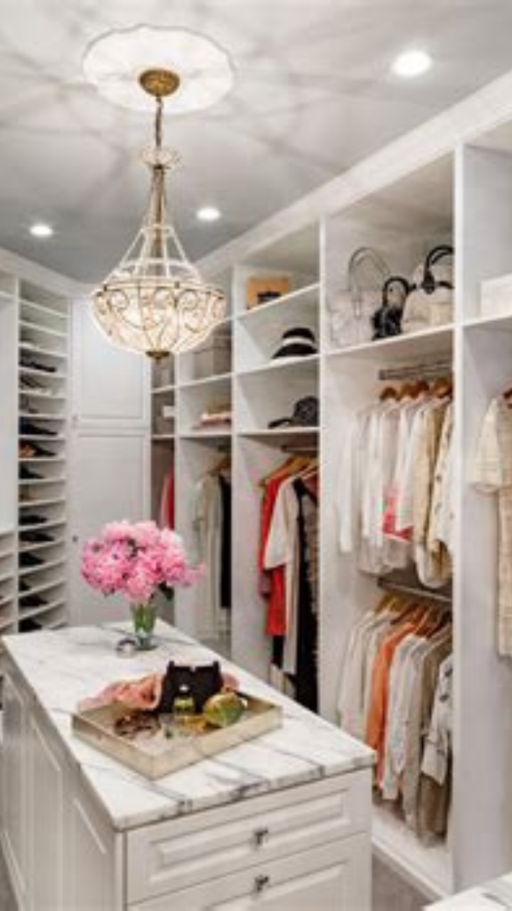 Elegant walk-in closet pin via lessloom.com titled "Choose Essentials That Earn Their Place in Your Closet" for a women's fashion Capsule Wardrobe. Features customized white built-in shelving, an elegant crystal chandelier, a marble center island with pink flowers, and neatly hung neutral tops and dresses. Ideal visual reference for quiet luxury closet organization, Scandinavian minimalist wardrobe storage, and decluttering your functional clothing space.