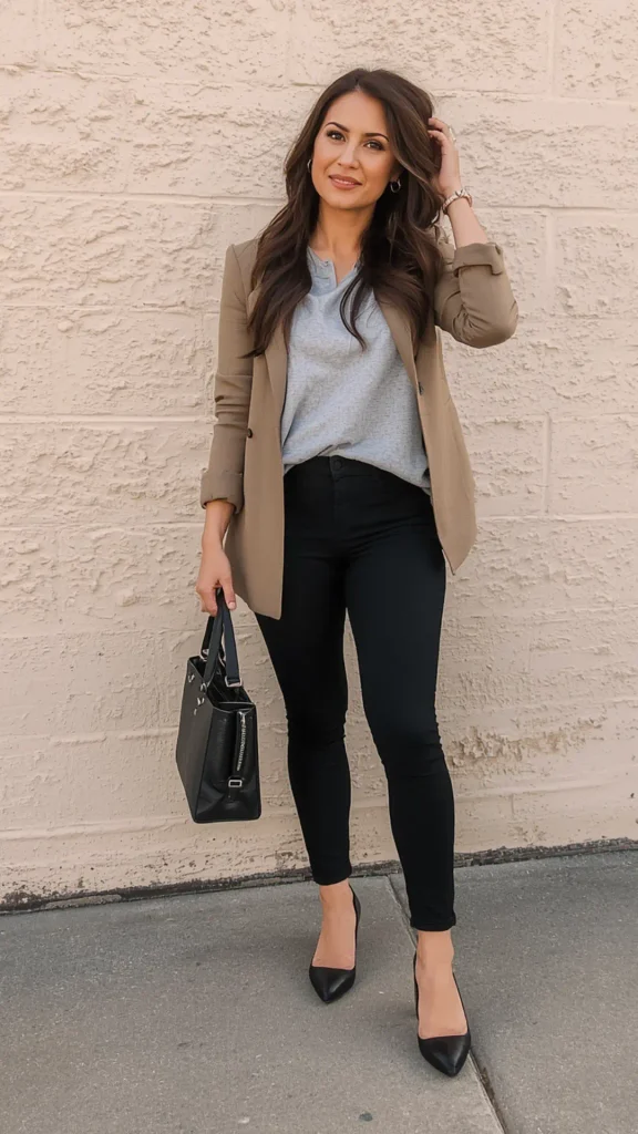 Full-length portrait of a woman standing by a beige wall wearing a beige blazer, a soft grey top, black fitted trousers, and black pointed heels for a women's fashion Capsule Wardrobe. Carrying a structured black leather tote bag. Ideal visual reference for neutral color palette mixing, quiet luxury workwear, Scandinavian minimalist office outfits, and timeless daily outfit formulas.